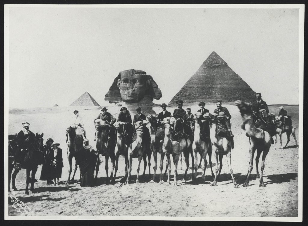 Photograph of riders on camels with the Sphinx and pyramids in the background.
